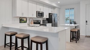 A modern kitchen with white cabinetry, a central island with stools, stainless steel appliances, and a large window providing natural light. - Heritage Landing Golf Club 30 night minimum 2 Bedroom Condo 2 Condo (Punta Gorda)
