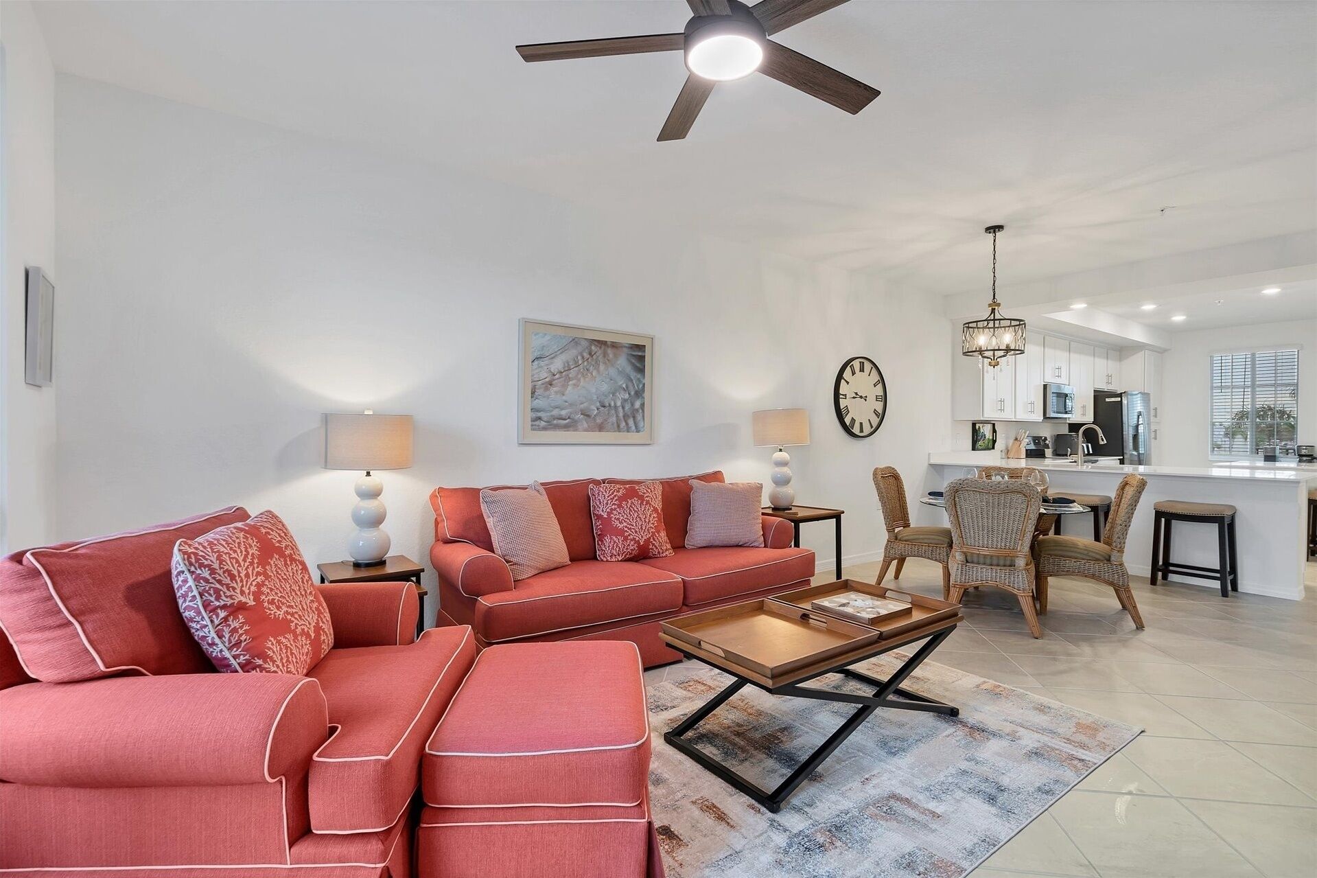 A living room with a sofa set, a coffee table, and a dining area with chairs. There is a ceiling fan, a clock, and a kitchen in the background.