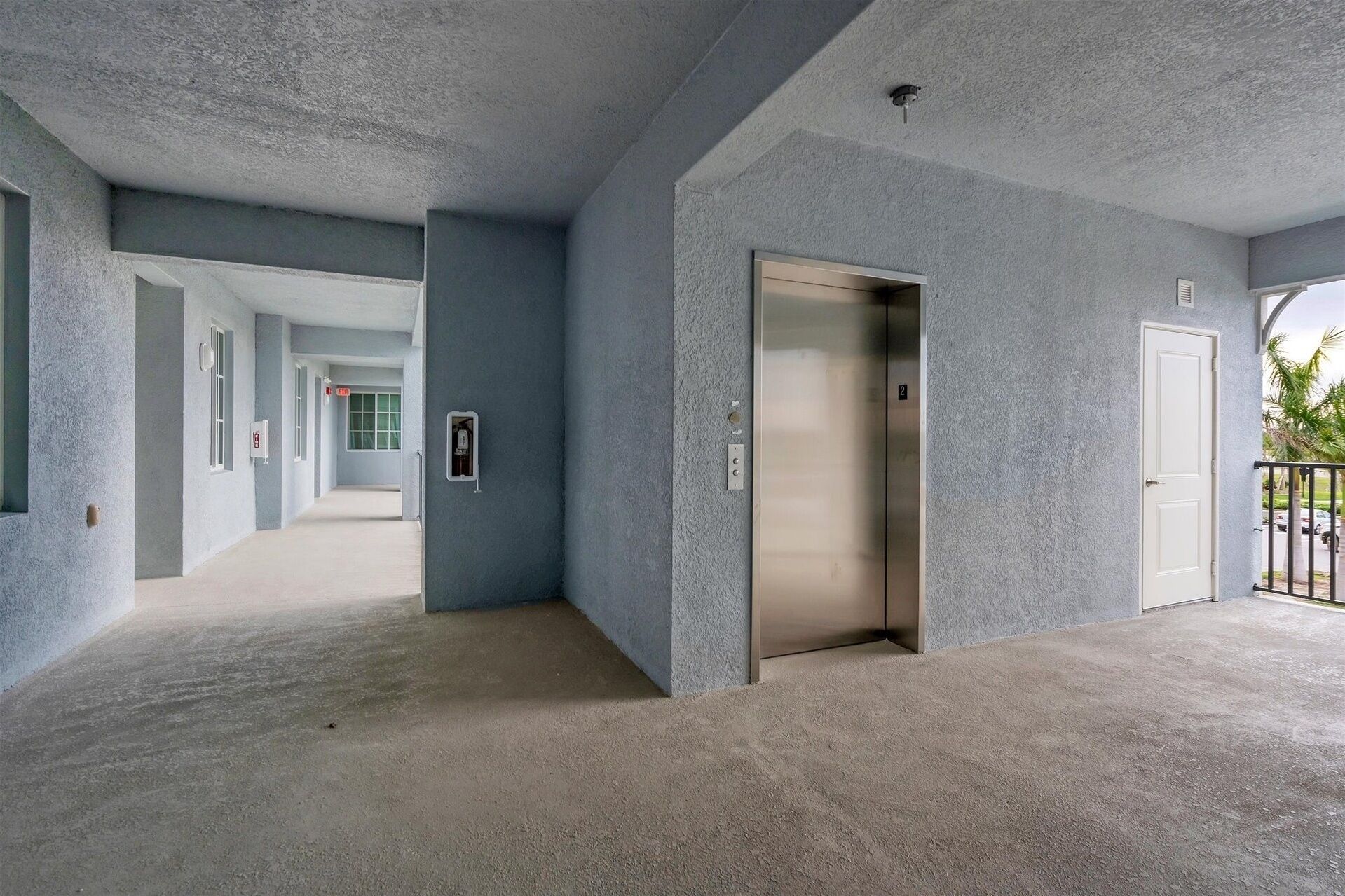 A hallway with a concrete floor and walls, an elevator, and a door leading to an outdoor area with a railing.