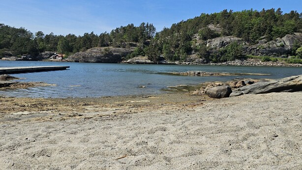 A beach with a dock extending into calm waters, surrounded by rocky terrain and dense forest. - Villa with sauna, 650 m to the beach | SE09135 (Varekil)