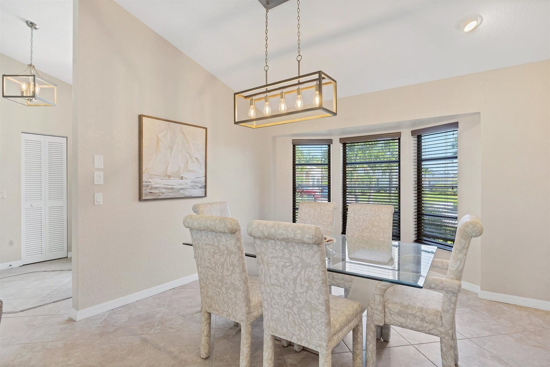 A dining area with a glass table and four chairs, a painting on the wall, and a ceiling light fixture.