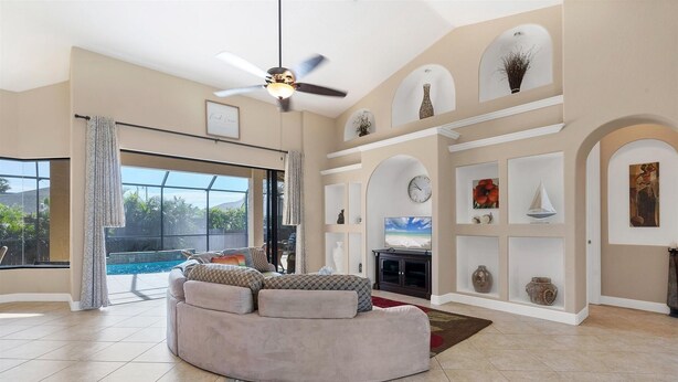 Un salón amplio con un sofá curvo, un ventilador de techo, un televisor grande y una piscina visible a través de las puertas de cristal.