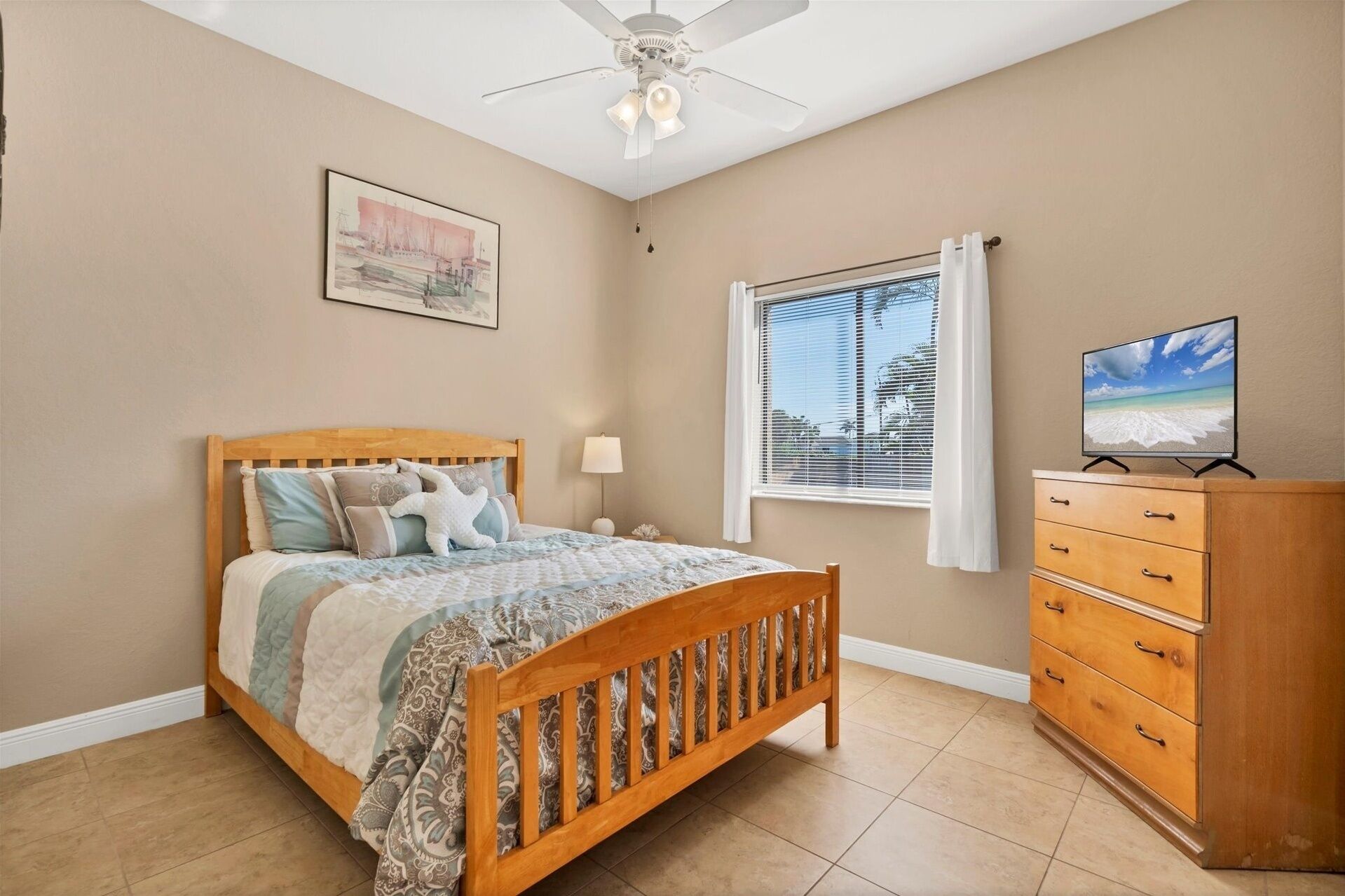 A bedroom with a wooden bed, a nightstand, a dresser, a ceiling fan, a window with blinds, and a painting on the wall.