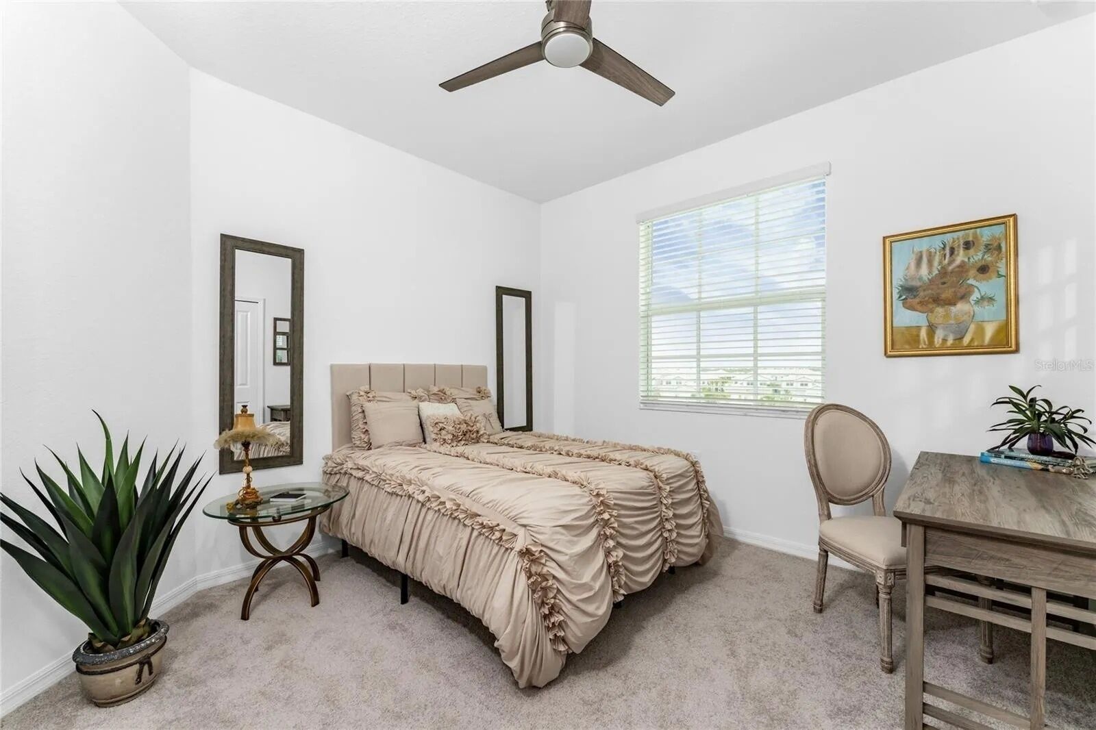 Une chambre à coucher comprenant un lit, un bureau, une chaise, un ventilateur de plafond, une fenêtre et un tableau.