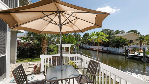 A balcony with a table and chairs, a large umbrella, and a view of a canal with boats and houses. - Lemon Bay Waterfront Condo W Patio, 5 Miles To Gulf Beaches 2 Bedroom Condo (Englewood)