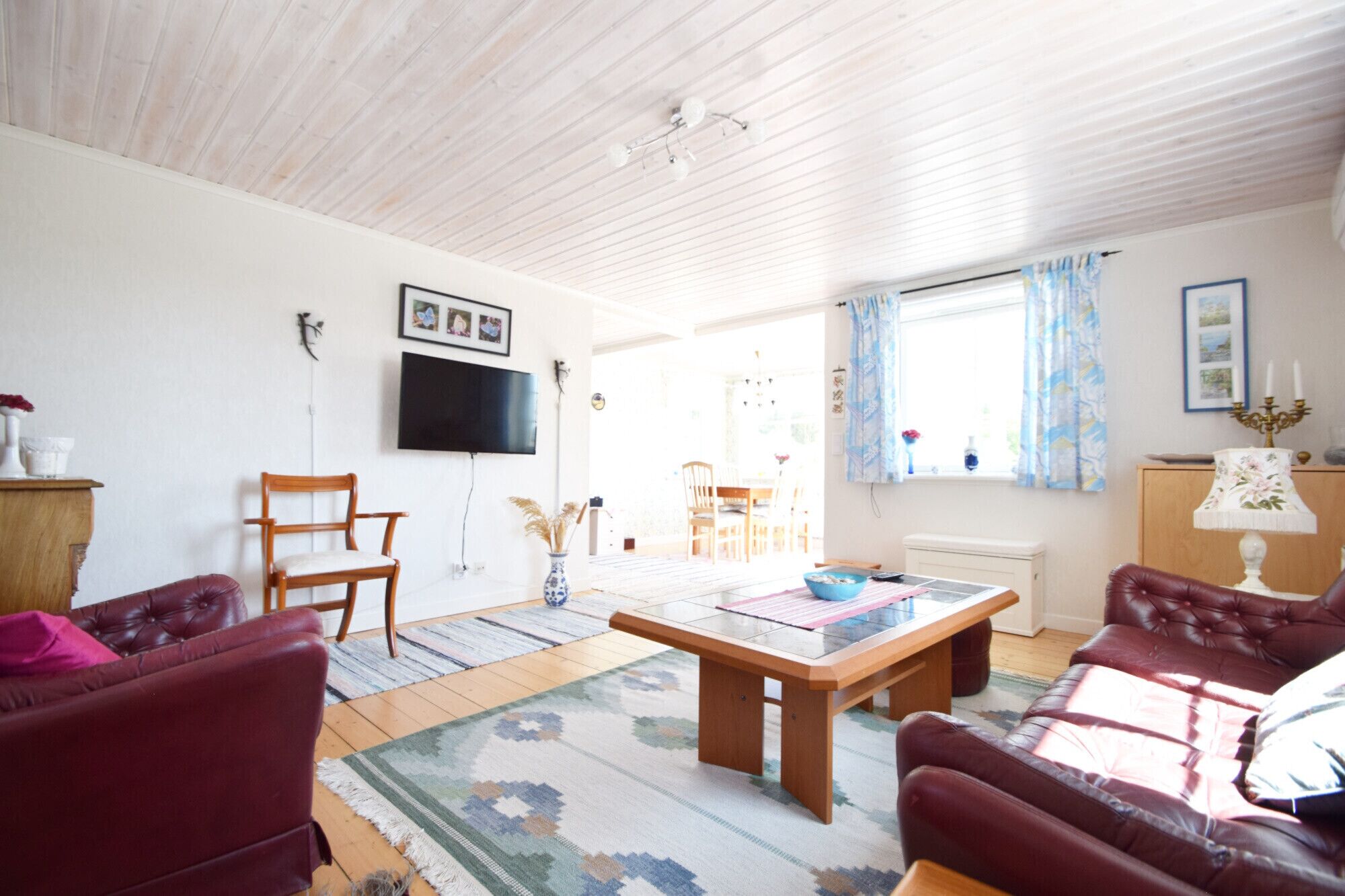 Un salon avec un plafond en bois, un téléviseur fixé au mur, un coin repas avec une table et des chaises, et une fenêtre avec des rideaux.