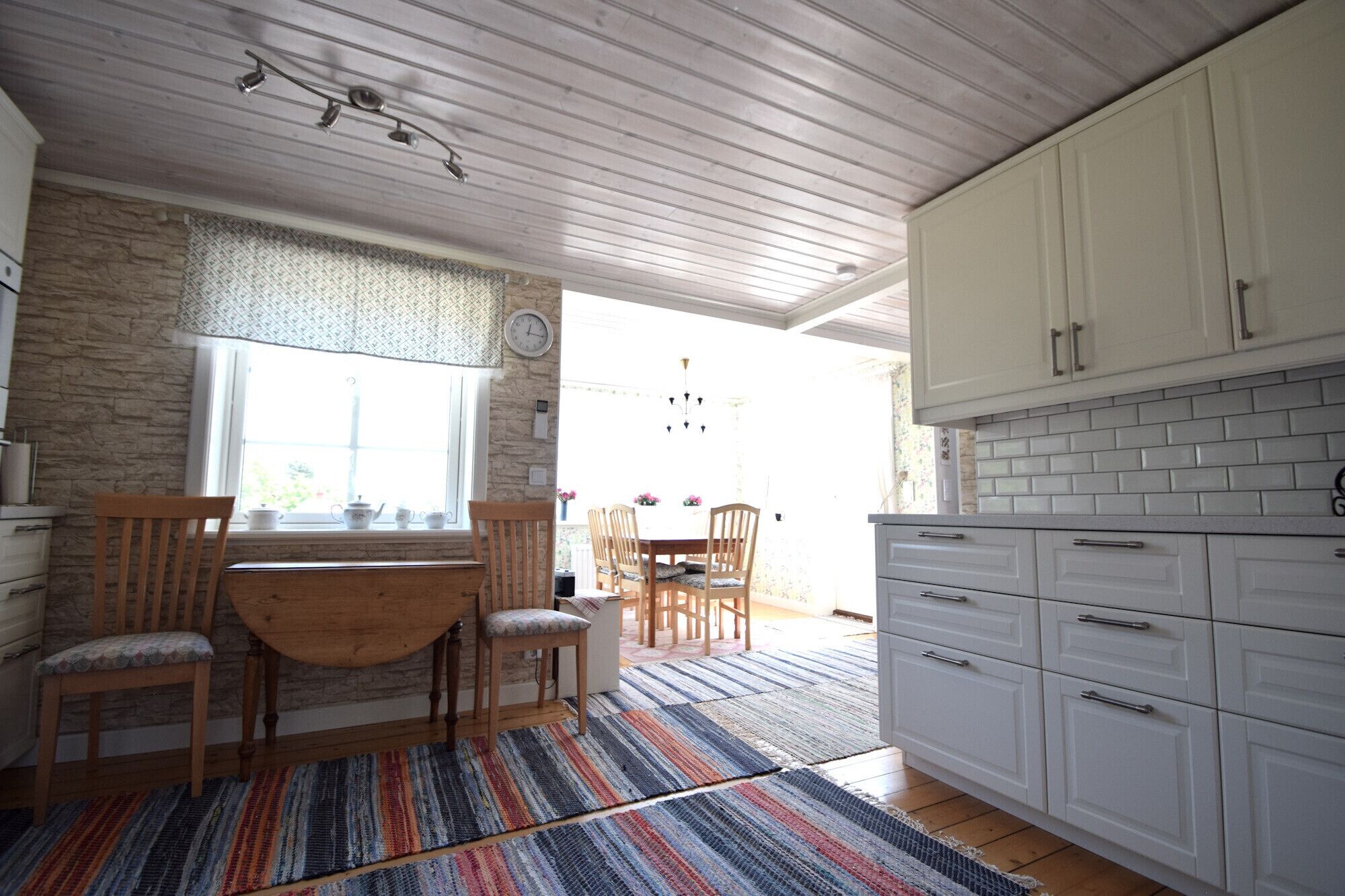 A kitchen with white cabinets, a dining area with a wooden table and chairs, a patterned rug, and a stone wall.