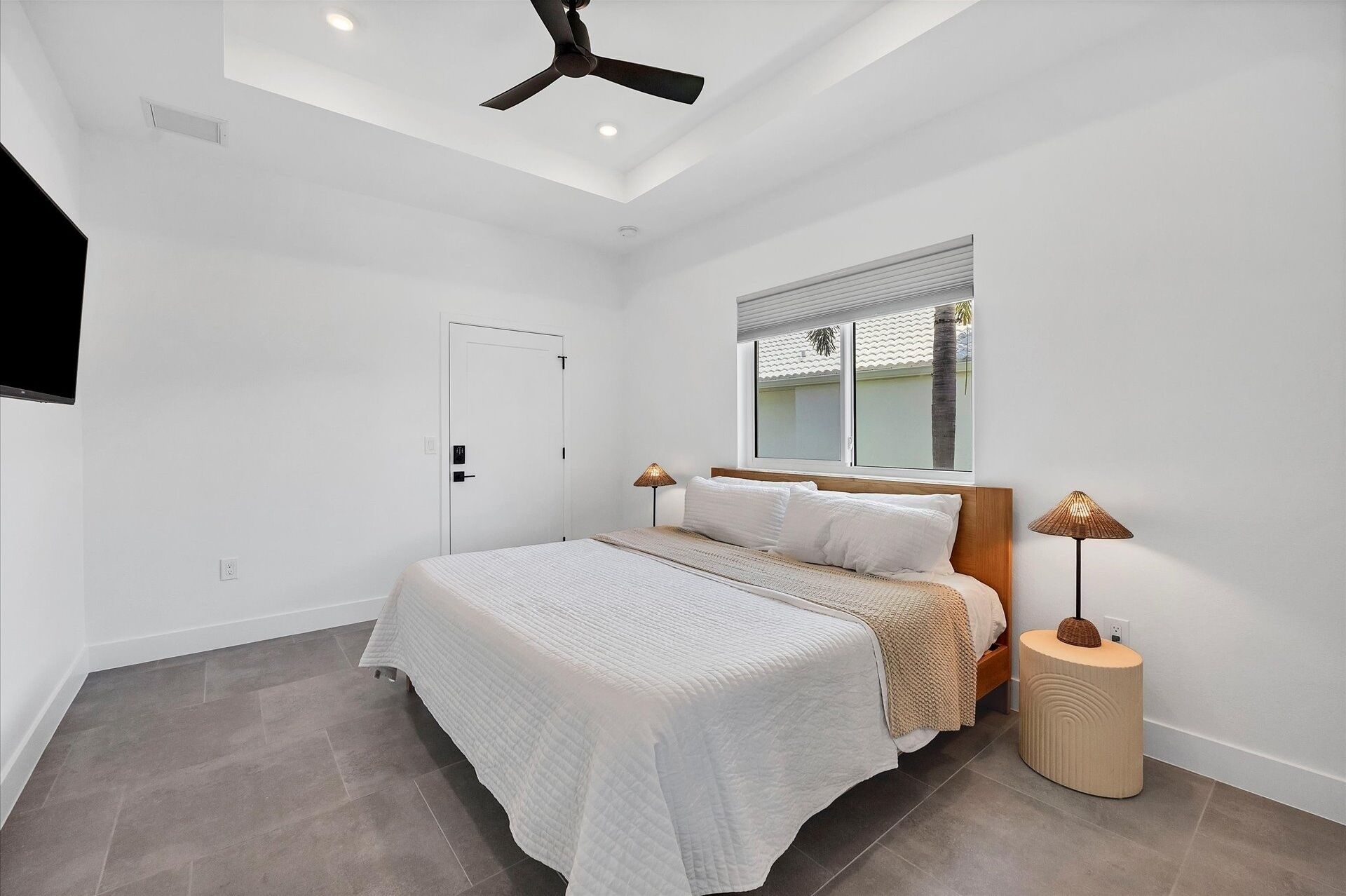 A bedroom with a bed, a ceiling fan, a window with blinds, a bedside lamp, and a door.