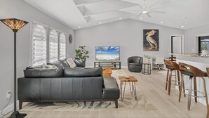 A modern living room with a grey sofa, a dining area with bar stools, a ceiling fan, and a large TV screen. - Punta Gorda Isles 3-Bedroom Canal-front Pool Home with Dock 3 Home (Punta Gorda)