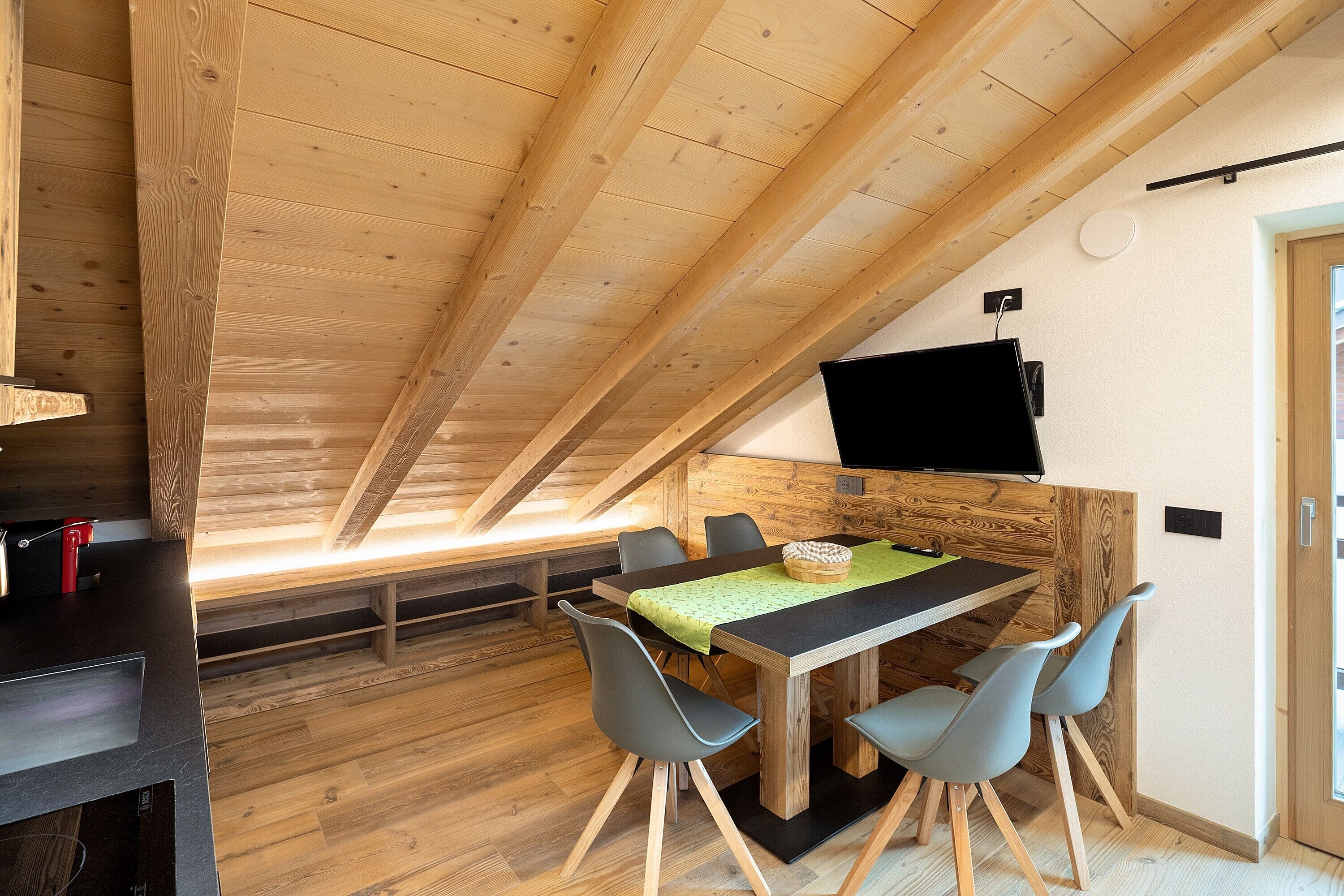 A dining area with a wooden table, four chairs, and a television mounted on the wall.