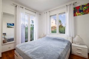 Ein Schlafzimmer mit einem Bett, zwei Nachttischen, einem Fenster mit Vorhängen, einem Gemälde und Blick ins Grüne.