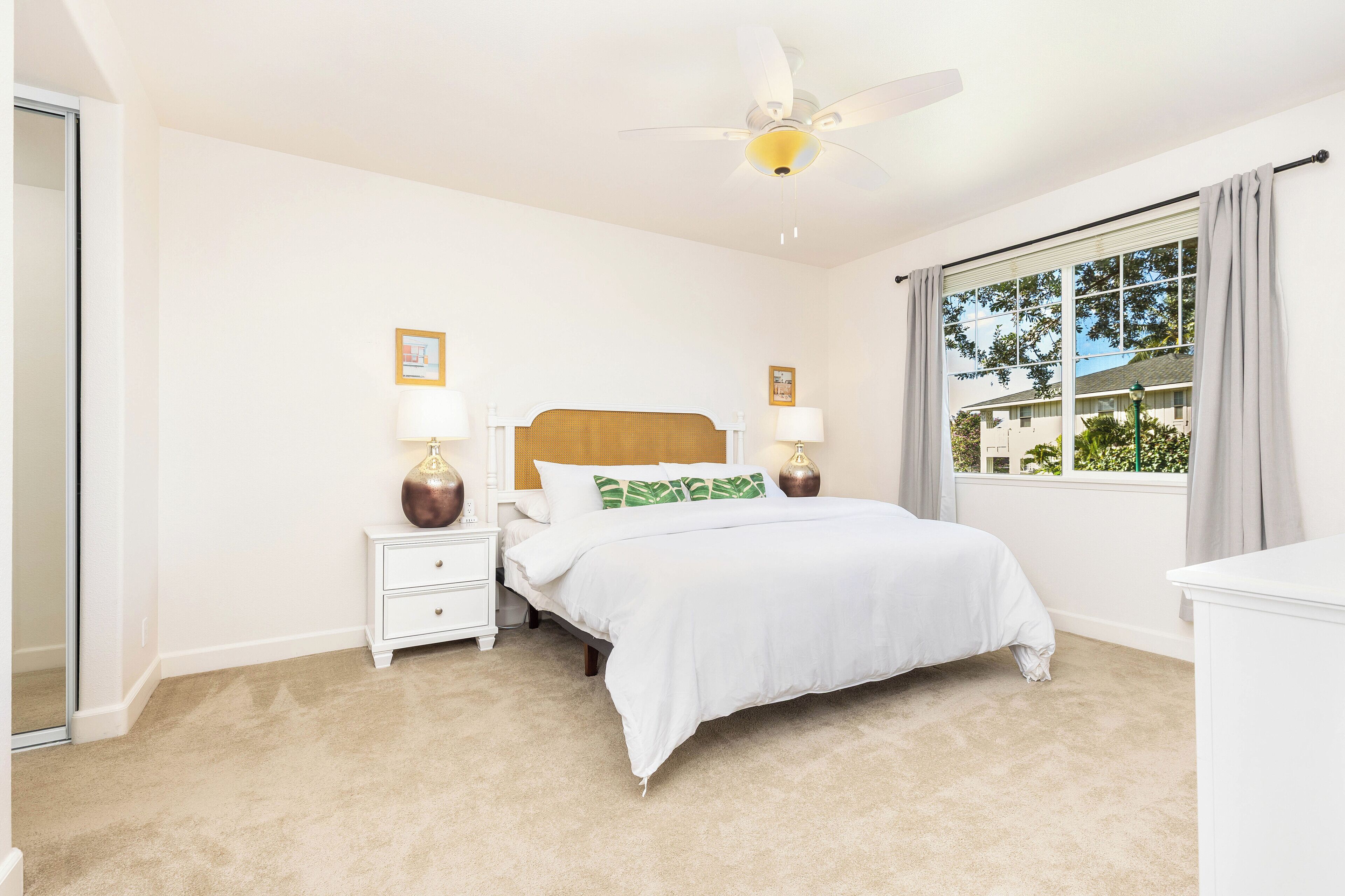 A bedroom with a single bed, two bedside tables with lamps, a ceiling fan, and a large window with curtains.