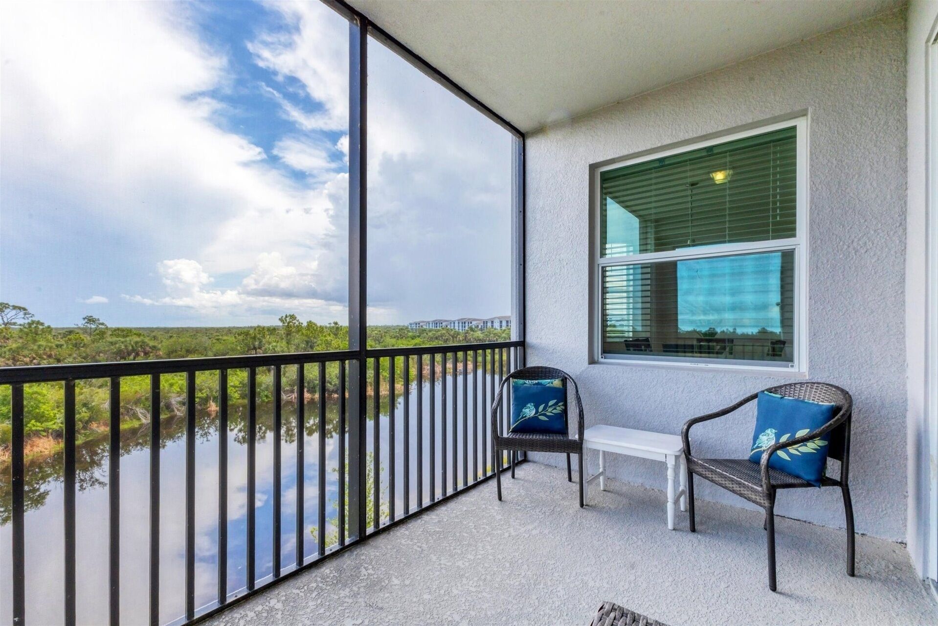 Un balcon donnant sur un plan d’eau et sur la verdure, une fenêtre avec des stores et deux fauteuils en osier avec des coussins.
