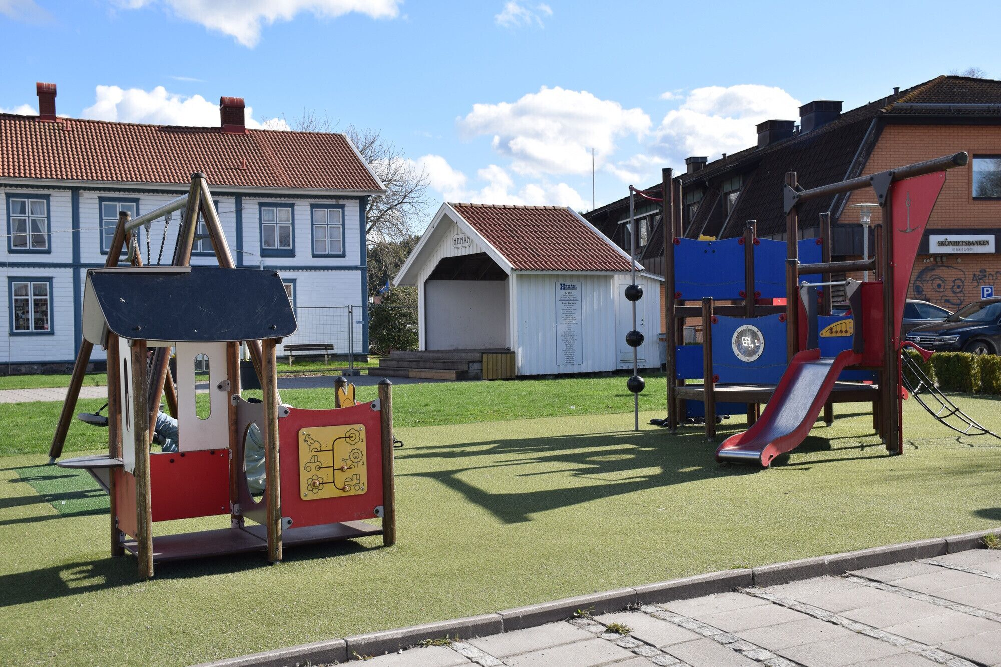 Ein Spielplatz mit Rutsche und Klettergerüsten, ein kleines weißes Gebäude mit der Aufschrift "HENAN" und im Hintergrund ein Wohngebäude mit rotem Ziegeldach.