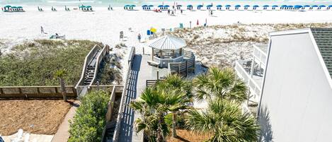 A beach view with a wooden boardwalk leading to a gazebo, palm trees, and beach umbrellas.