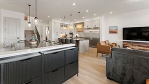 Et moderne køkken med en central ø, rustfrie hårde hvidevarer, hængende lamper og et spiseområde med stole.