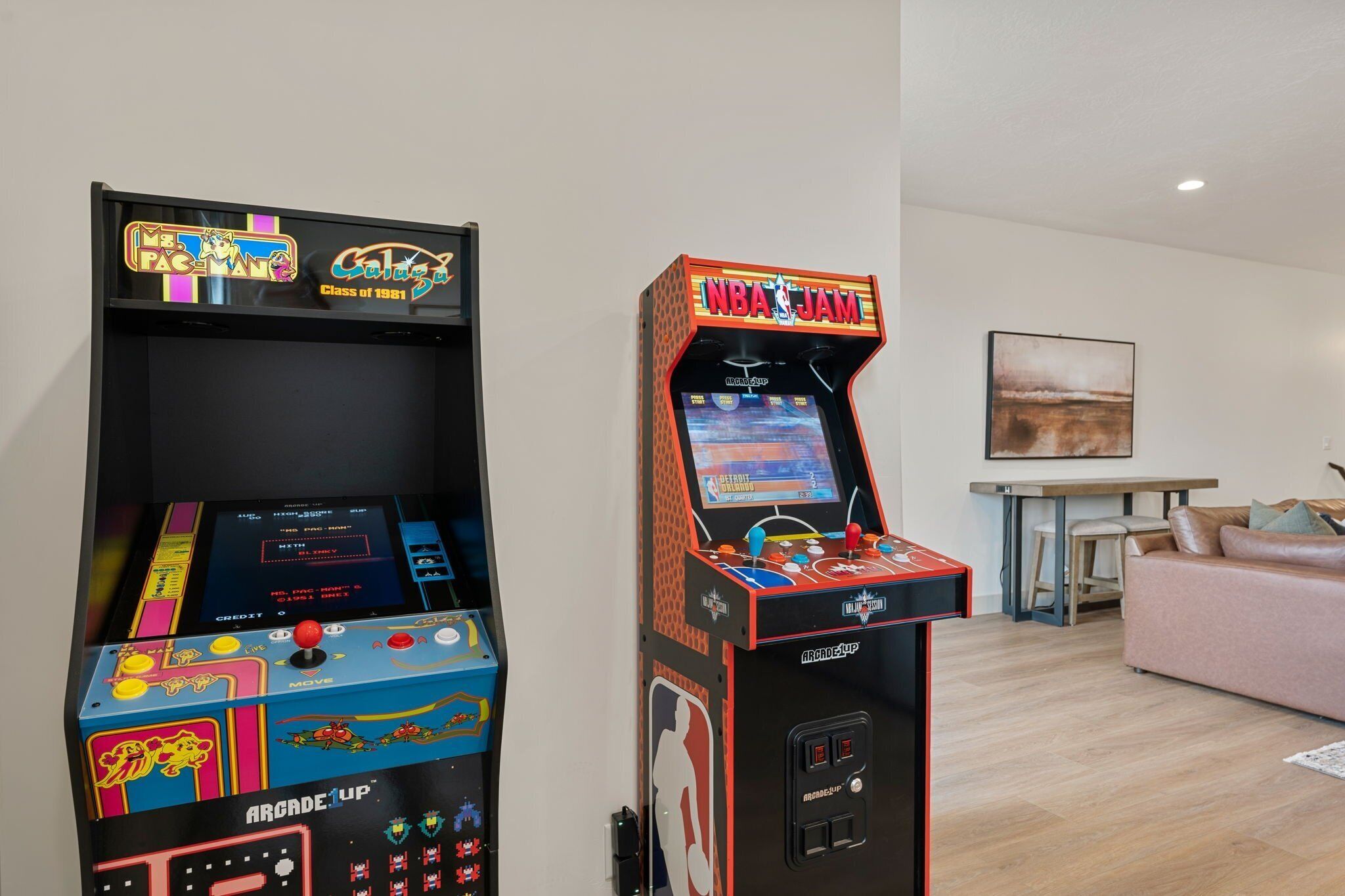 Twee speelautomaten, een Pac-Man en een NBA Jam, in een kamer met een bank en een schilderij aan de muur.