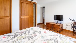 Une chambre avec des armoires en bois, un lit recouvert d’une couvre-lit à motifs floraux, un téléviseur posé sur une commode en bois et une chaise.
