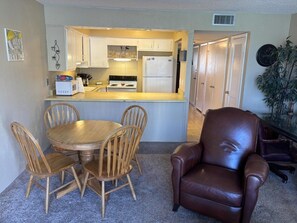 A compact kitchen with white cabinets, a refrigerator, and a dining area with a wooden table and chairs. - Vantage Pointe 138 - Condo Across The Street In Daytona Beach 1 Bedroom Condo (Daytona Beach)