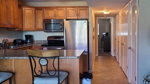 A kitchen with wooden cabinets, a stainless steel refrigerator, a microwave, and a coffee maker. There is a chair with a cushion and a table with a lamp. - Vantage Pointe #222 1 Bedroom Condo (Daytona Beach)