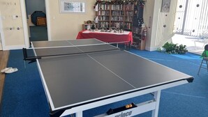 A ping pong table in a room with a bookshelf, a table with decorations, and a door leading to another room. - Vantage Pointe #222 1 Bedroom Condo (Daytona Beach)