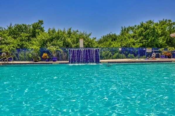 A swimming pool with a blue fence, surrounded by lush greenery. - Kid Friendly Condo With Pool, Basketball, Bocce, Hot Tub, And More! 3 Bedroom Condo (New Smyrna Beach)