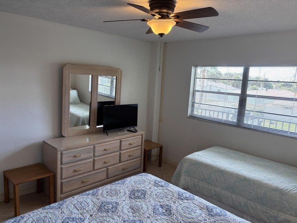 Une chambre à coucher comprenant un lit, une commode, un téléviseur, un ventilateur de plafond et une fenêtre avec des stores.