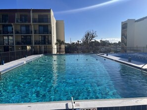Piscina com água azul cristalina, cercada por grade, ao lado de um edifício com varandas.