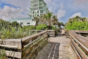 A wooden boardwalk leading to a multi-story building with balconies, surrounded by lush greenery and palm trees. - Oceanfront Condo With Beautiful Views 2 Bedroom Condo (New Smyrna Beach)