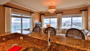 A kitchen with granite countertops, a stainless steel sink, and a chandelier. There are two large windows with curtains, and a balcony with a view of the city. - Oceanfront Condo With Beautiful Views 2 Bedroom Condo (New Smyrna Beach)