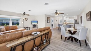 Un vaste espace de vie comprenant un grand canapé, une table basse en bois et une table à manger avec des chaises. On peut également apercevoir des ventilateurs de plafond, un téléviseur et une cuisine au fond de la pièce.