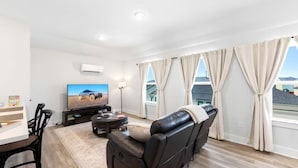 Ein modernes Wohnzimmer mit Flachbildfernseher, einer Ledersofa, einem Esstisch und einem großen Fenster mit Vorhängen.