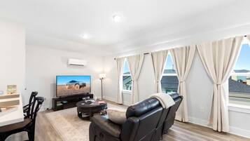 Ein modernes Wohnzimmer mit Flachbildfernseher, einer Ledersofa, einem Esstisch und einem großen Fenster mit Vorhängen.