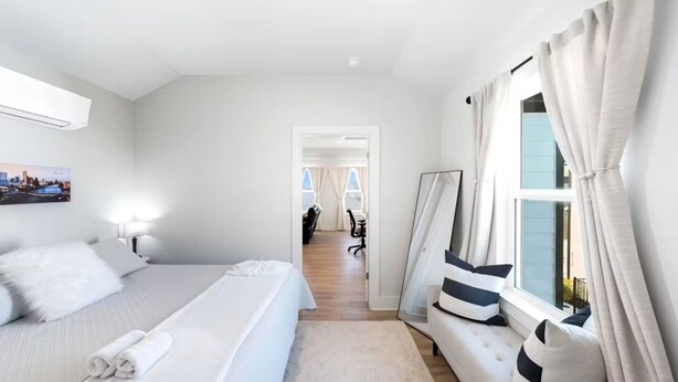 Ein Hotelzimmer mit einem Bett, einem Schreibtisch, einem Stuhl, einem fenster mit Vorhängen und einem Gang zu einem anderen Zimmer.