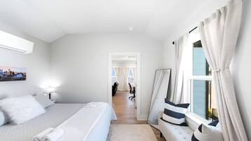 Ein Hotelzimmer mit einem Bett, einem Schreibtisch, einem Stuhl, einem fenster mit Vorhängen und einem Gang zu einem anderen Zimmer.