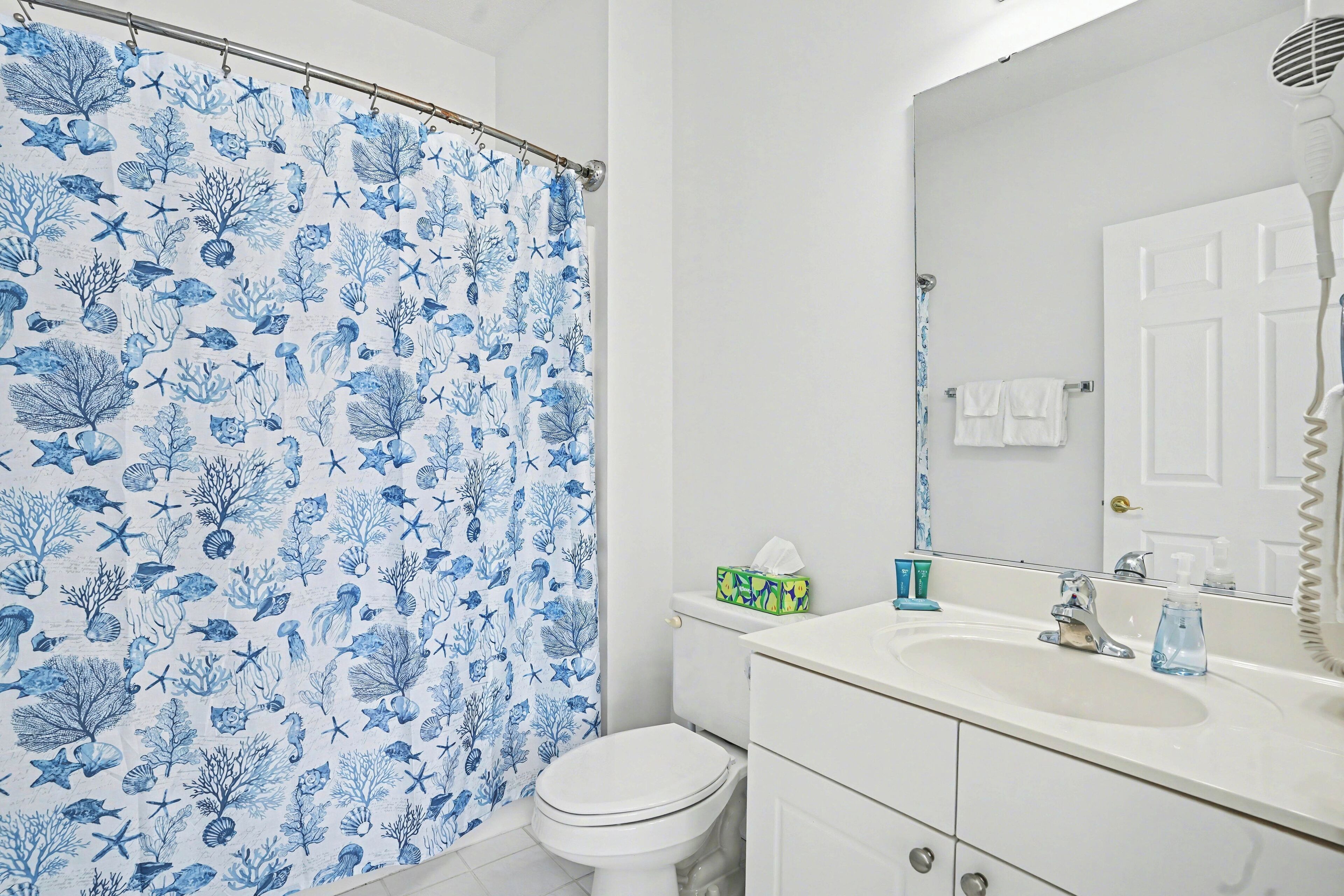 Ein Badezimmer mit Duschvorhang mit blauem Meereslebewesen-Muster, einer weißen Toilette, einem Waschbecken mit Spiegel und Pflegeprodukten auf der Ablagefläche.