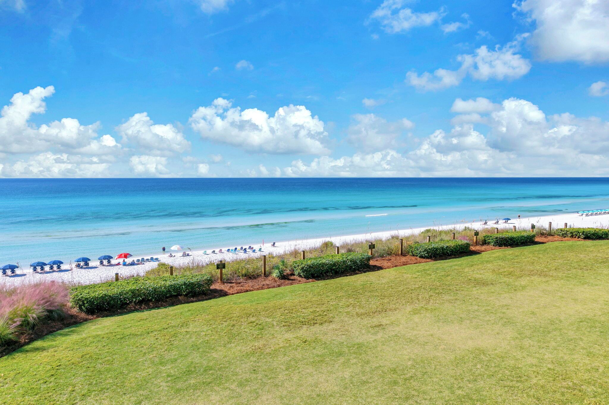 A beach with a grassy foreground, a sandy beach, and a clear blue sky with clouds.
