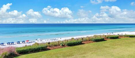 A beach with a grassy foreground, a sandy beach, and a clear blue sky with clouds.