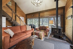 A cozy cabin interior with a wooden staircase, a sofa, a chair, a fireplace, and large windows. - Cluster Cabin 13 1 Bedroom Cabin (Bend)