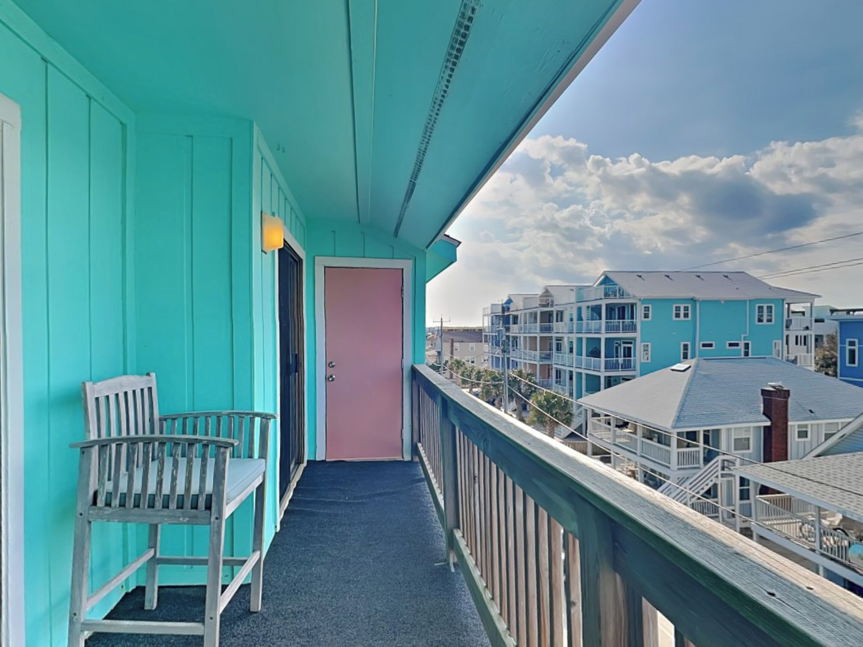 Un balcon turquoise avec une chaise en bois et une porte, donnant sur une rangée de bâtiments bleus.