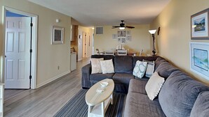 Ein Wohnzimmer mit einem grauen Ecksofa, einem kleinen runden Tisch und einem Deckenventilator. Eine Tür führt in einen anderen Raum, im Hintergrund ist ein Essbereich zu sehen.