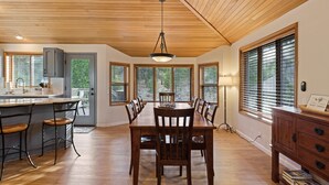 A spacious dining area with a wooden table and chairs, a kitchen area with a bar, and large windows offering views of the outdoors. - Tan Oak 2 3 Bedroom Home (Bend)