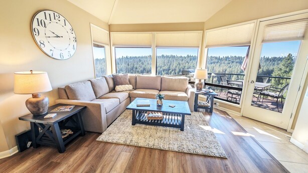 A living room with a sectional sofa, a coffee table, a clock, and large windows offering a forest view. - Mt Bachelor Village 424 A Sr 2 Bedroom Condo (Bend)