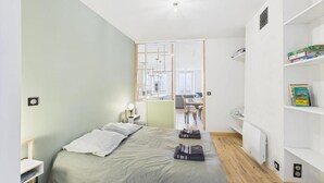 A modern bedroom with a bed, bedside tables, a radiator, and a view into the living area. - Cosy apartment in the center of town - Philotée 2 (Clermont-Ferrand)