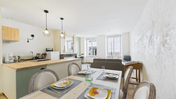 A dining area with a table set for four, a kitchen in the background, and a view of the city through large windows. - Cosy apartment in the center of town - Philotée 2 (Clermont-Ferrand)