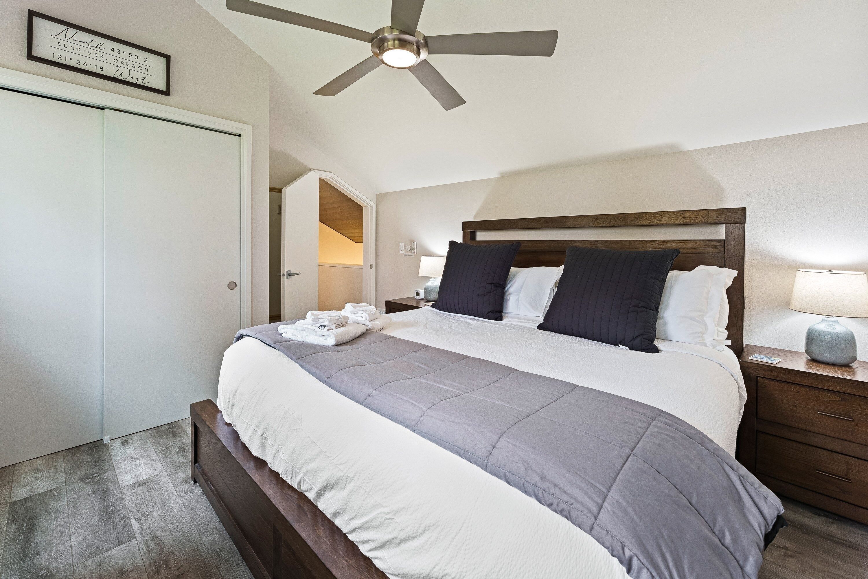 A bedroom with a bed, a ceiling fan, a nightstand, a lamp, and a framed sign on the wall.