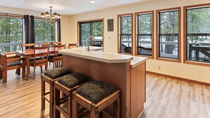 A modern kitchen with a central island, wooden dining table, and large windows offering views of the outdoors. - Yellow Pine 10 5 Bedroom Home (Bend)