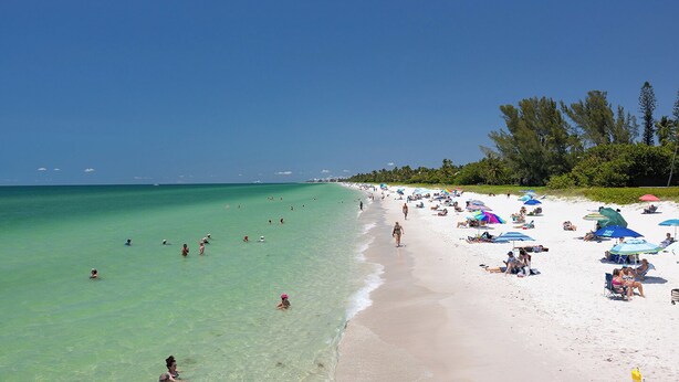Een strand met helder turquoise water, wit zand en mensen die zwemmen en zonnebaden.