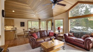 A spacious living area with a large brown leather sofa, a wooden coffee table, and a dining table with chairs. There are large windows providing natural light and views of the outdoors. - Lark 4 2 Bedroom Home (Bend)