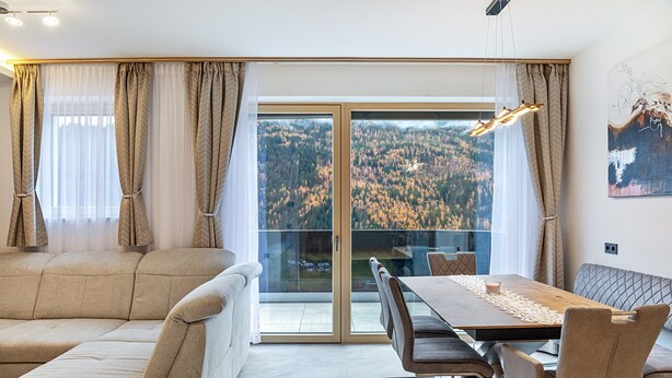 Ein modernes Wohnzimmer mit einer Couch, einem Esstisch, Stühlen und Blick durch eine Glastür auf ein bewaldetes Gebiet.